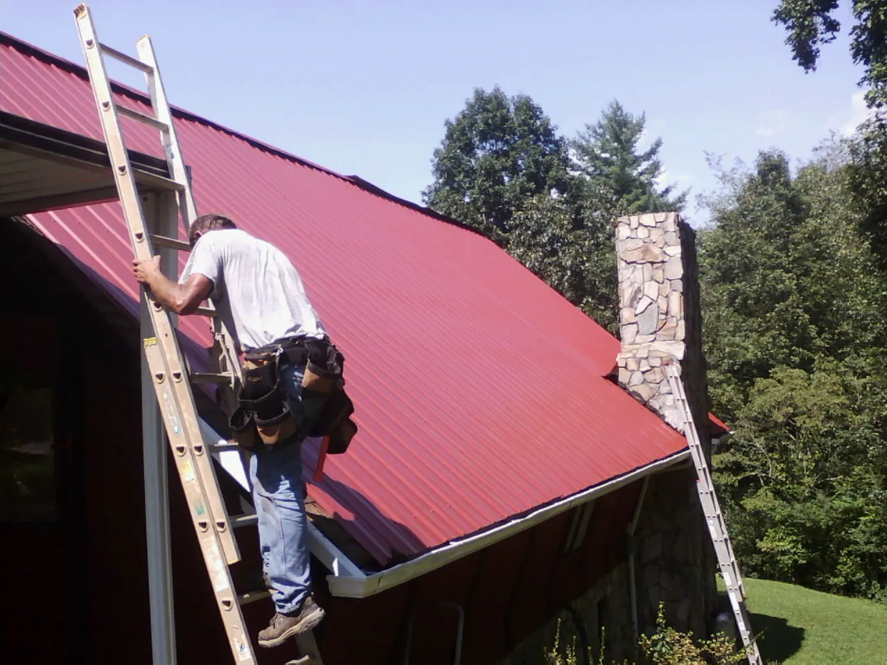 Roofer on a ladder beside a red metal roof and stone chimney — Soffit & Fascia Repair by Roofing Brothers in Dyke